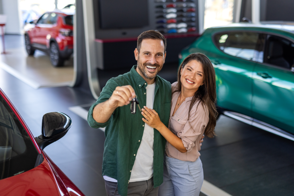 Happy couple holding car keys inside a dealership showroom, standing next to their new vehicle.